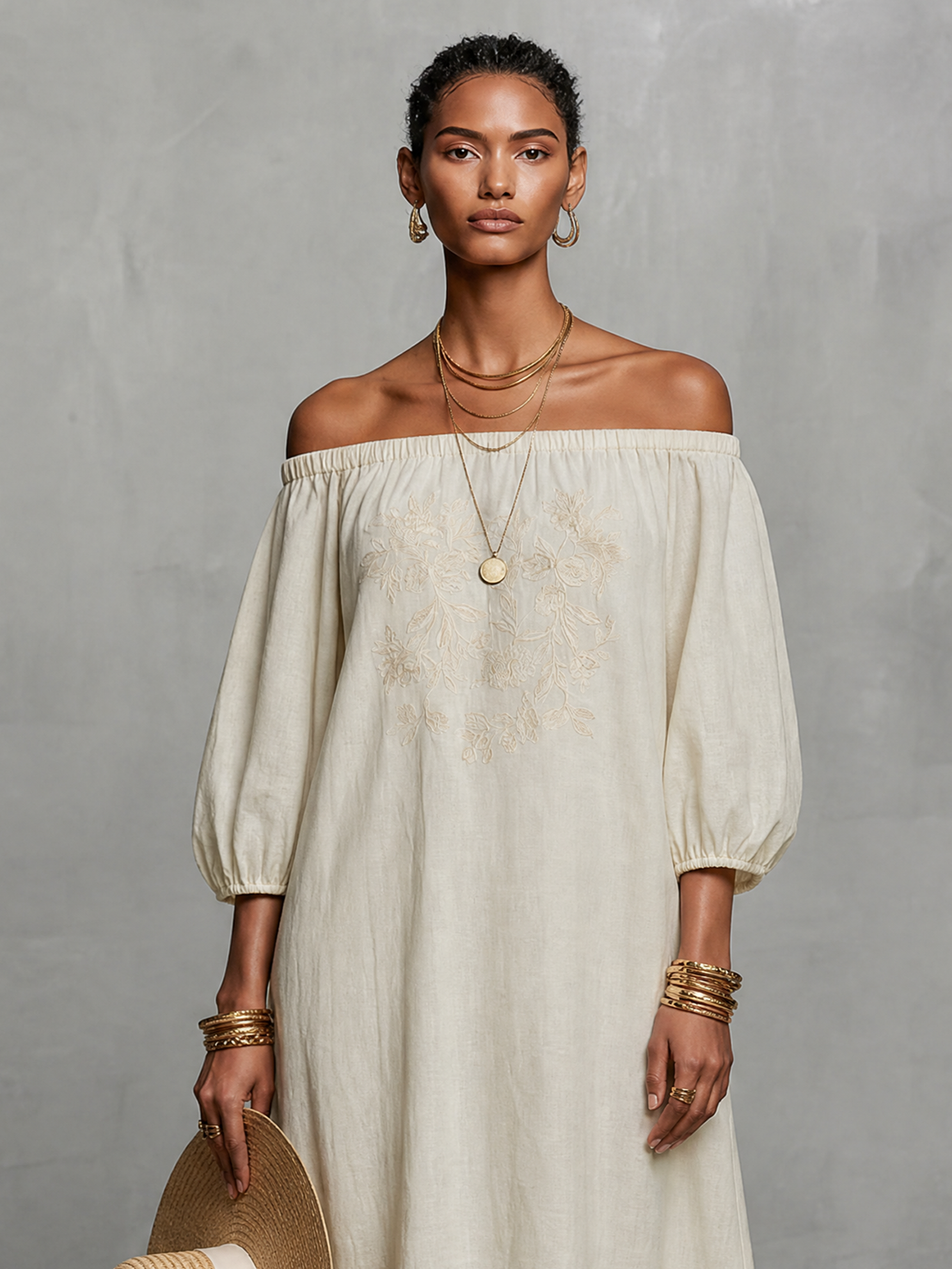 Black - A woman wearing an off-the-shoulder cream linen maxi dress with tonal chest embroidery and puff sleeves, holding a wide-brim straw hat.