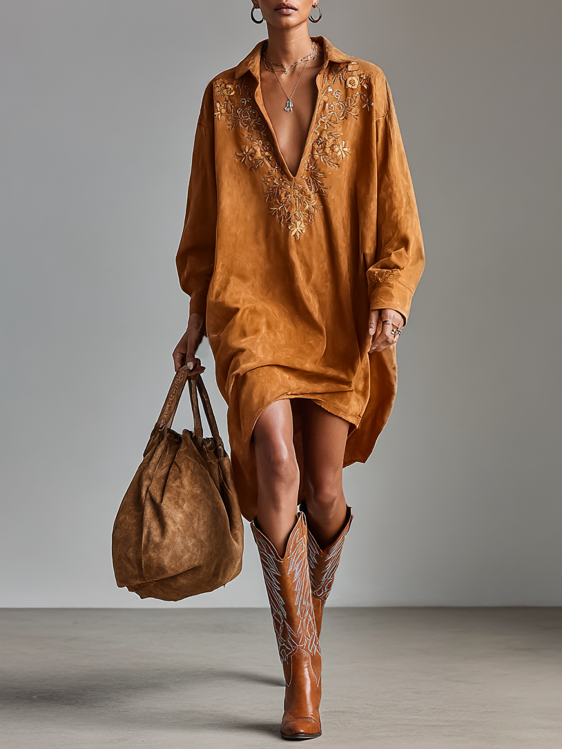 Brown - A model wearing a long-sleeved, ochre-coloured matte satin shift dress featuring a deep V-neckline with intricate floral embroidery, styled with knee-high tan cowboy boots.