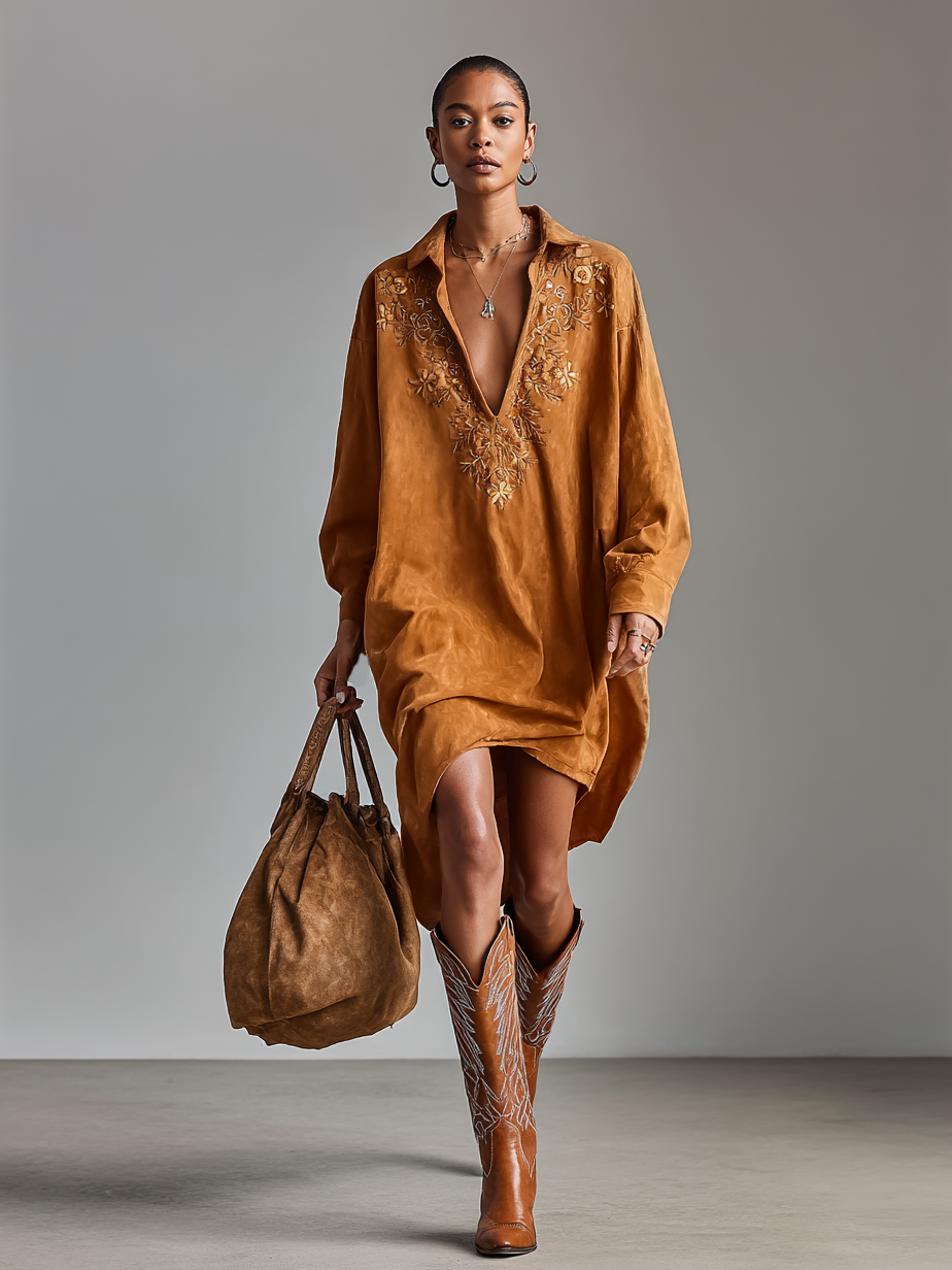 Brown - A model wearing a long-sleeved, ochre-coloured matte satin shift dress featuring a deep V-neckline with intricate floral embroidery, styled with knee-high tan cowboy boots.