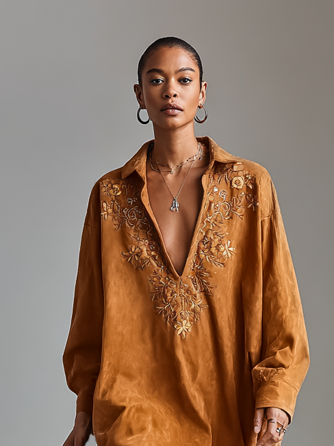 Brown - A model wearing a long-sleeved, ochre-coloured matte satin shift dress featuring a deep V-neckline with intricate floral embroidery, styled with knee-high tan cowboy boots.