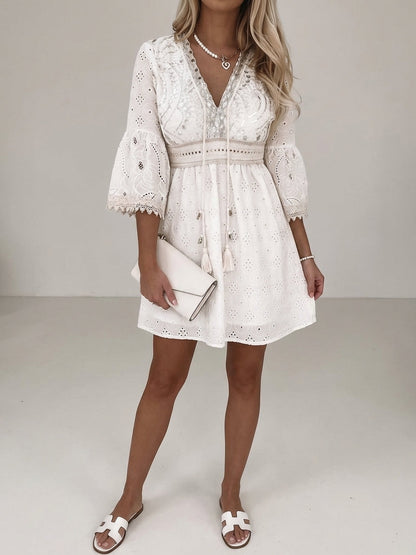 Beige - A woman wearing a cream-coloured eyelet lace mini dress with a V-neckline, tassel ties, three-quarter bell sleeves, and a tiered ruffled hem.