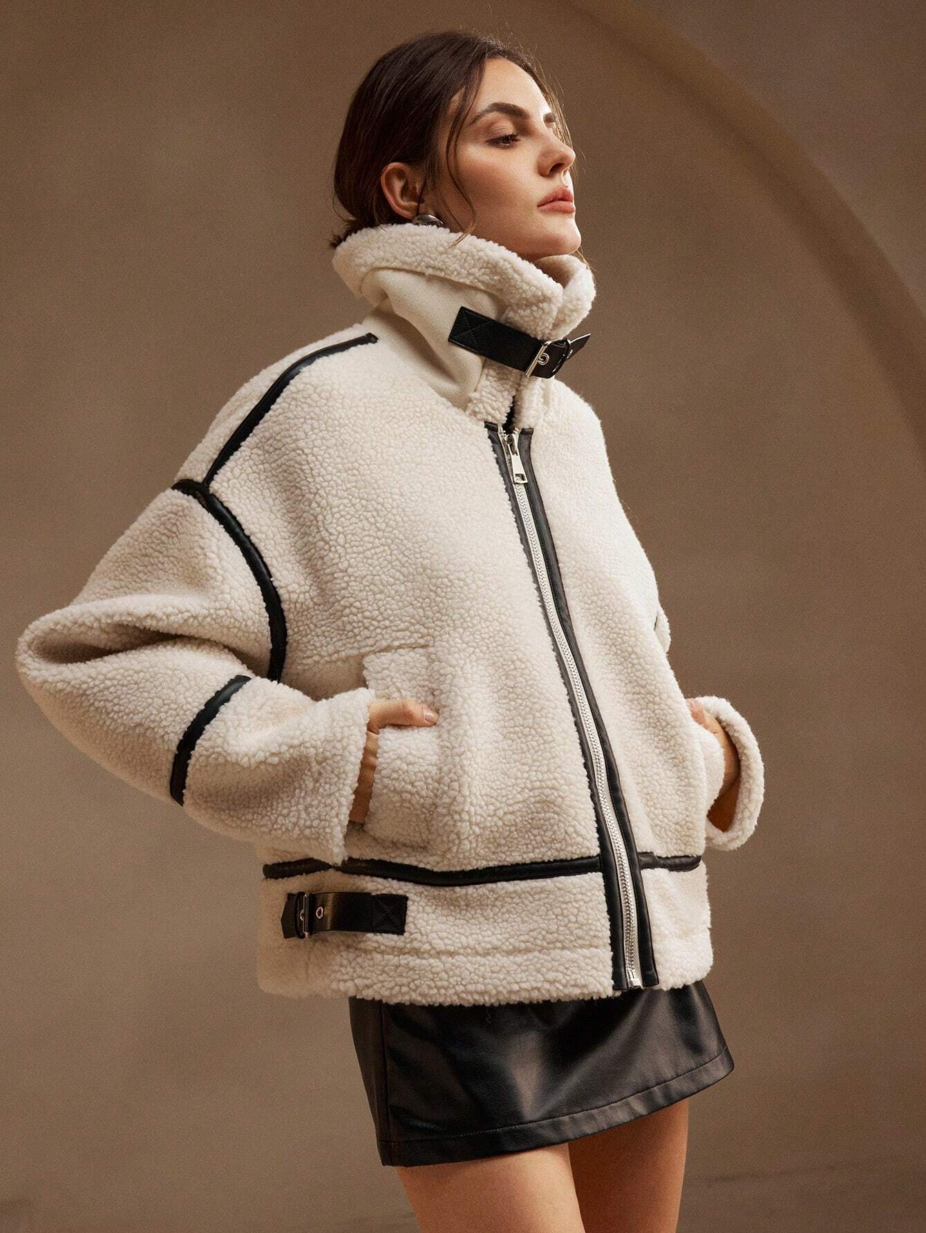 White - A woman lounging on a sofa wearing an oversized, cream-coloured faux shearling aviator jacket featuring striking black contrast taping, a front zipper, and a buckle detail on the collar.