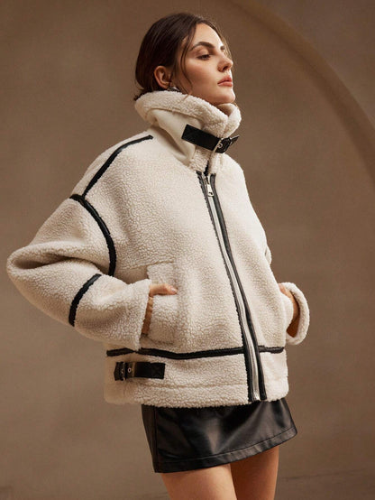 White - A woman lounging on a sofa wearing an oversized, cream-coloured faux shearling aviator jacket featuring striking black contrast taping, a front zipper, and a buckle detail on the collar.