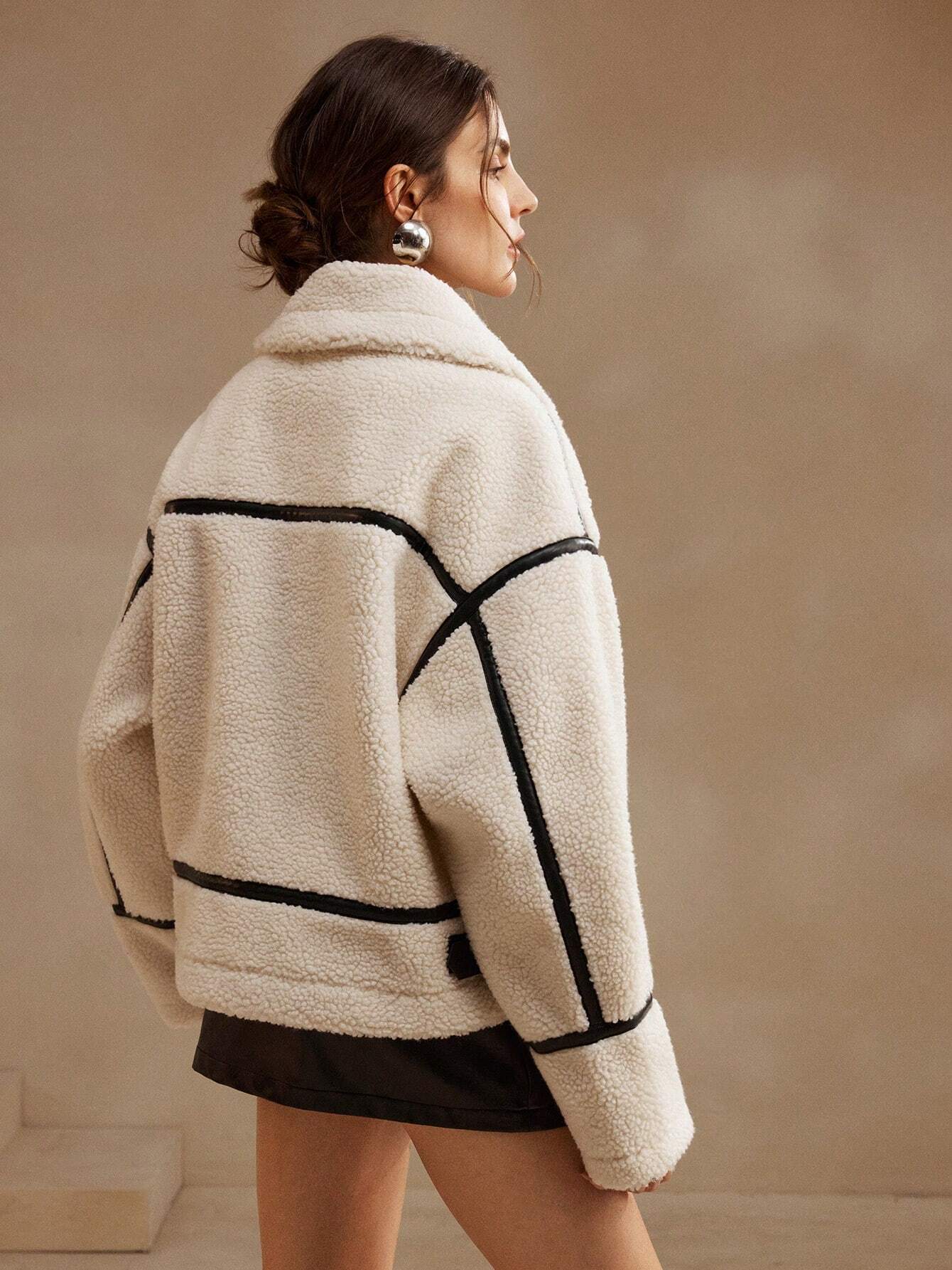 White - A woman lounging on a sofa wearing an oversized, cream-coloured faux shearling aviator jacket featuring striking black contrast taping, a front zipper, and a buckle detail on the collar.