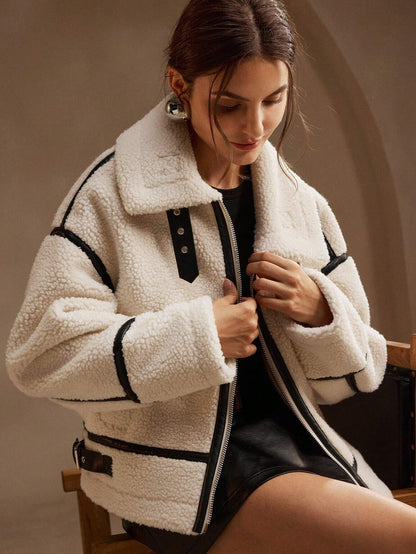 White - A woman lounging on a sofa wearing an oversized, cream-coloured faux shearling aviator jacket featuring striking black contrast taping, a front zipper, and a buckle detail on the collar.