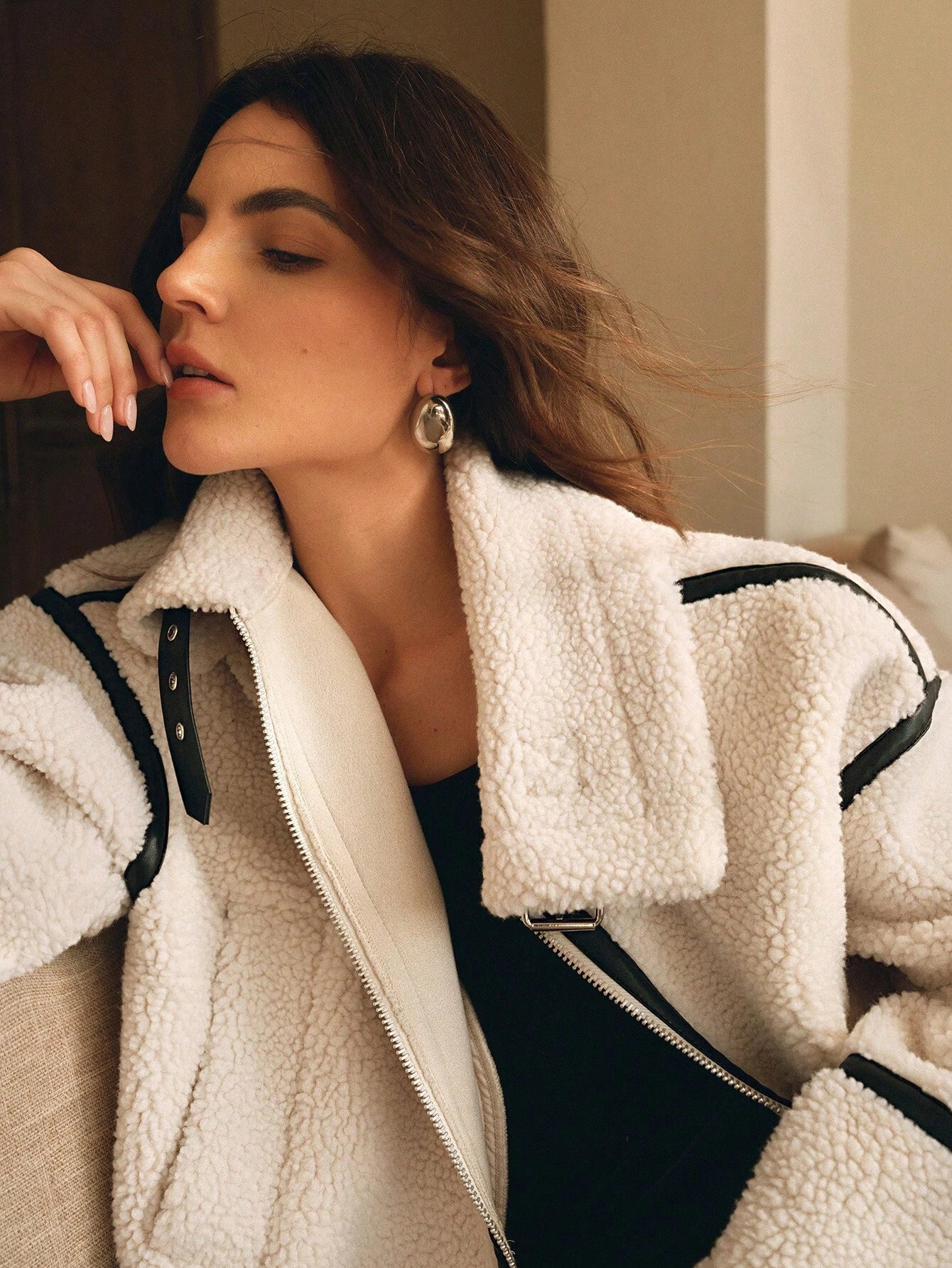 White - A woman lounging on a sofa wearing an oversized, cream-coloured faux shearling aviator jacket featuring striking black contrast taping, a front zipper, and a buckle detail on the collar.