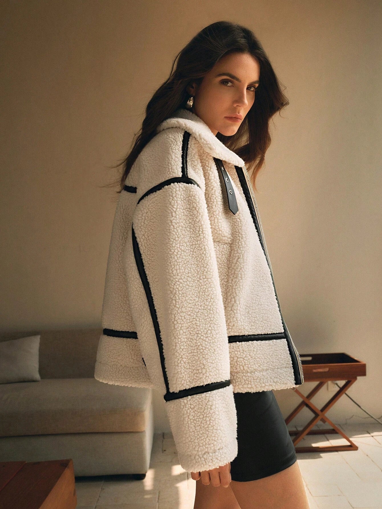 White - A woman lounging on a sofa wearing an oversized, cream-coloured faux shearling aviator jacket featuring striking black contrast taping, a front zipper, and a buckle detail on the collar.