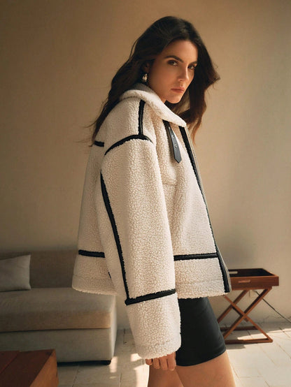 White - A woman lounging on a sofa wearing an oversized, cream-coloured faux shearling aviator jacket featuring striking black contrast taping, a front zipper, and a buckle detail on the collar.