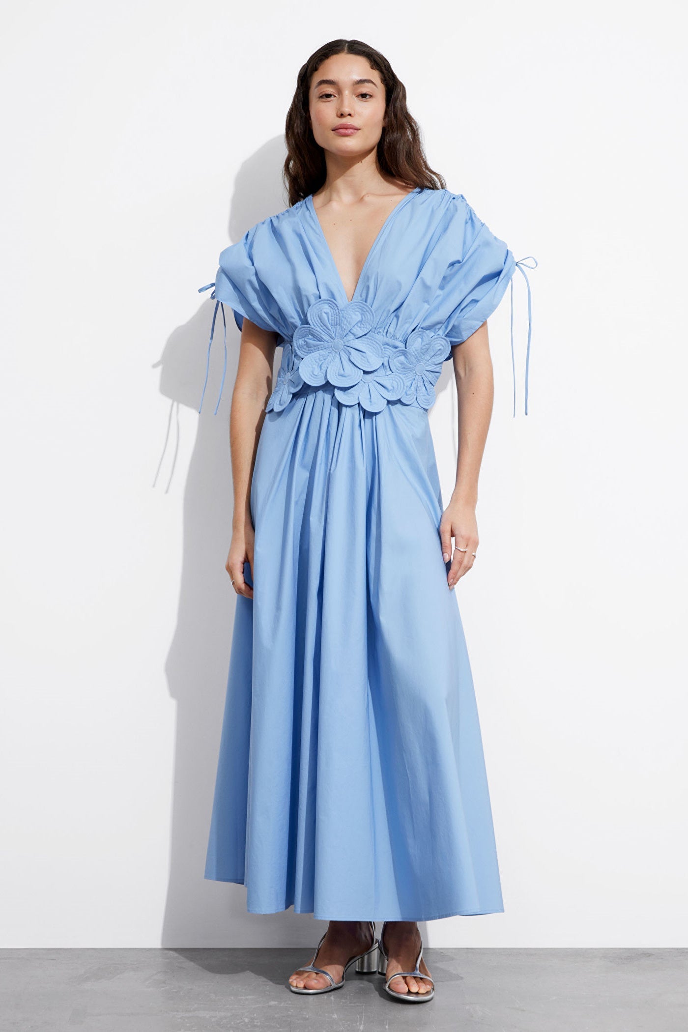 Blue - A woman wearing a light blue maxi dress featuring a deep V-neckline, voluminous short sleeves with tie details, and a prominent 3D floral applique at the waist.