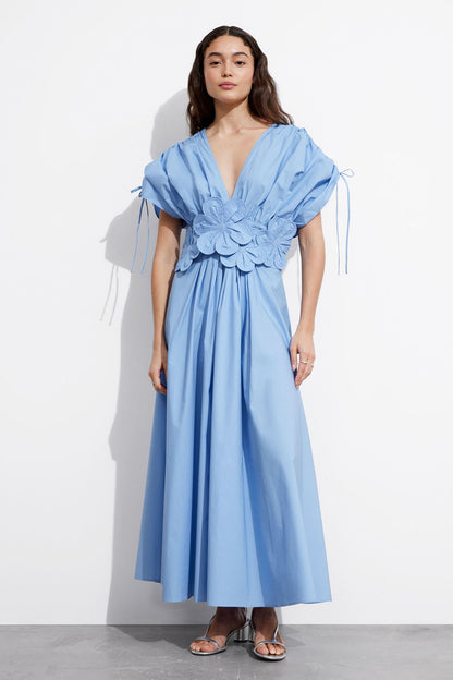 Blue - A woman wearing a light blue maxi dress featuring a deep V-neckline, voluminous short sleeves with tie details, and a prominent 3D floral applique at the waist.