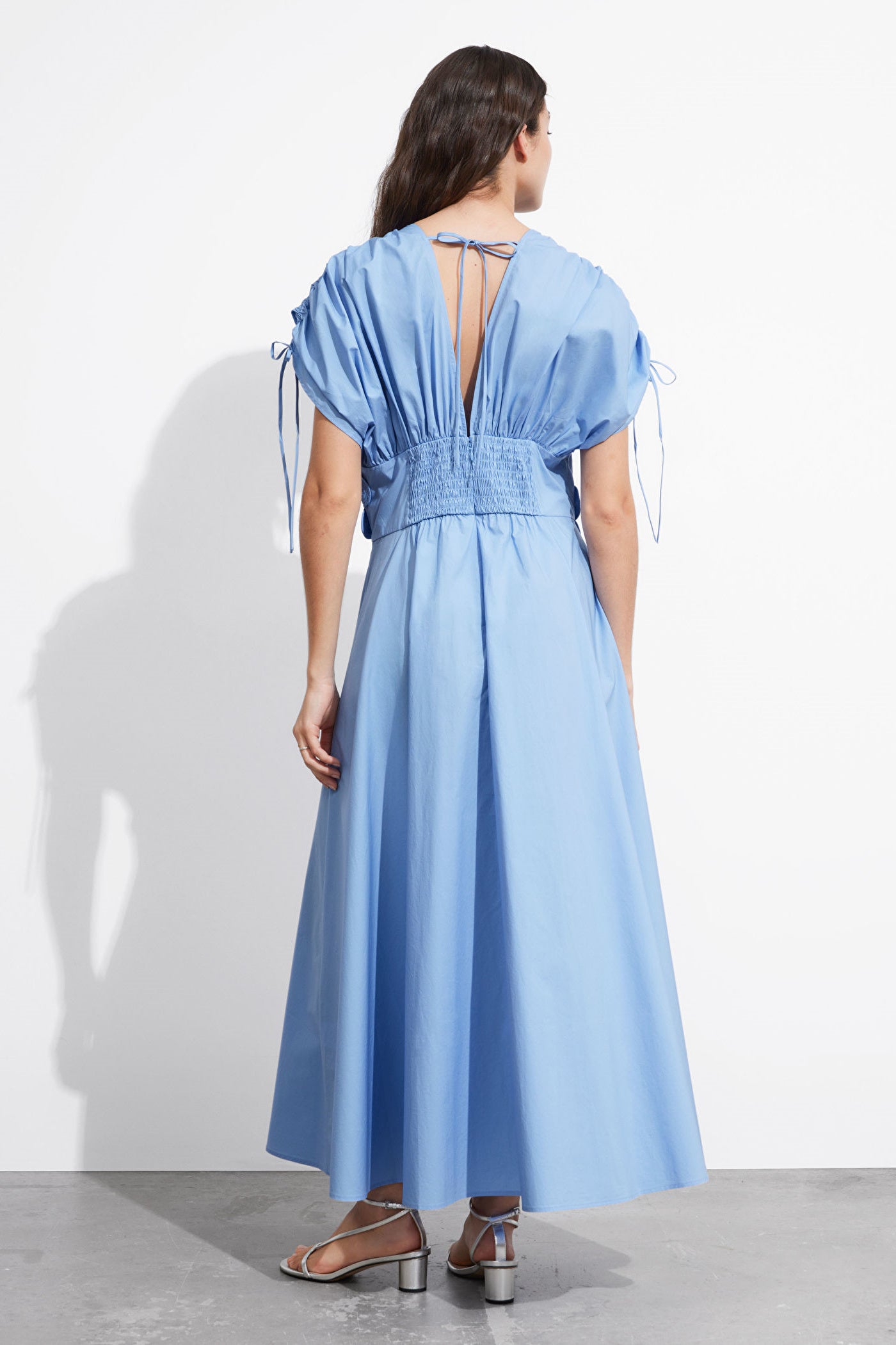 Blue - A woman wearing a light blue maxi dress featuring a deep V-neckline, voluminous short sleeves with tie details, and a prominent 3D floral applique at the waist.