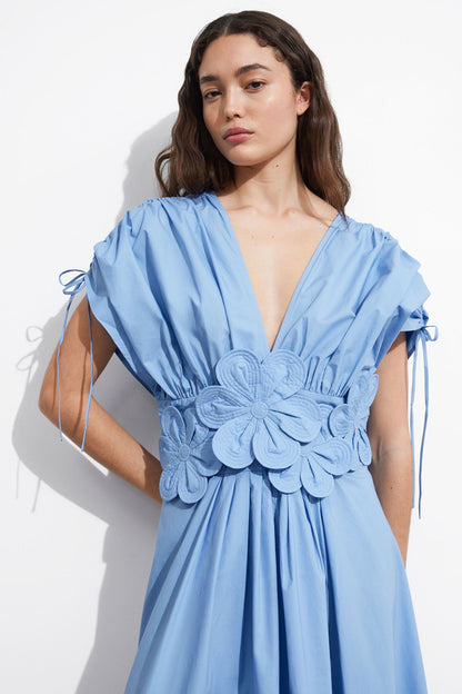Blue - A woman wearing a light blue maxi dress featuring a deep V-neckline, voluminous short sleeves with tie details, and a prominent 3D floral applique at the waist.