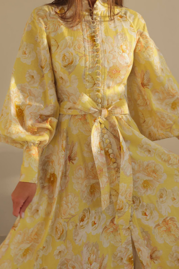 Yellow - A woman wearing a yellow and white floral print maxi dress with long balloon sleeves, a high neckline, button-front detailing, and a self-tie waist.