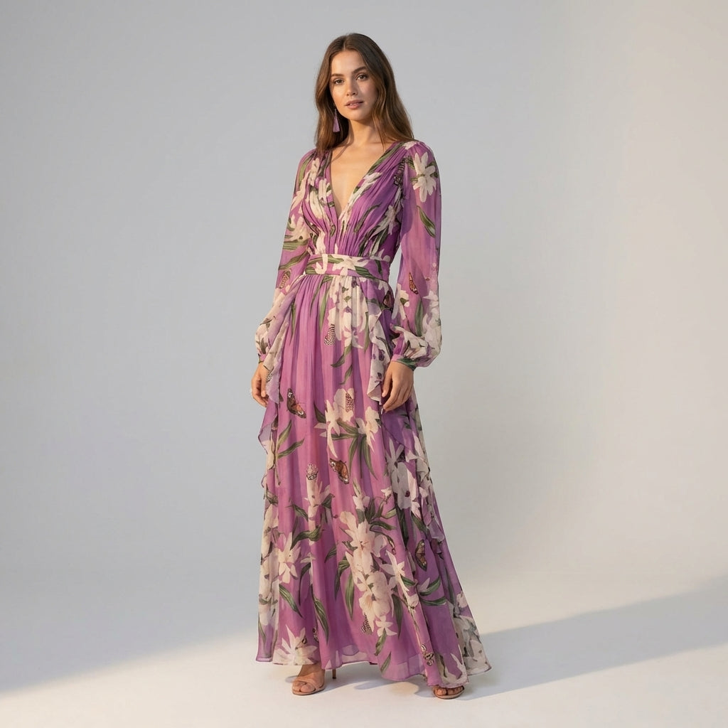 A woman stands wearing a floor-length lavender maxi dress made of sheer floral and butterfly print chiffon, featuring a deep V-neckline, pleated bodice, and long blouson sleeves.