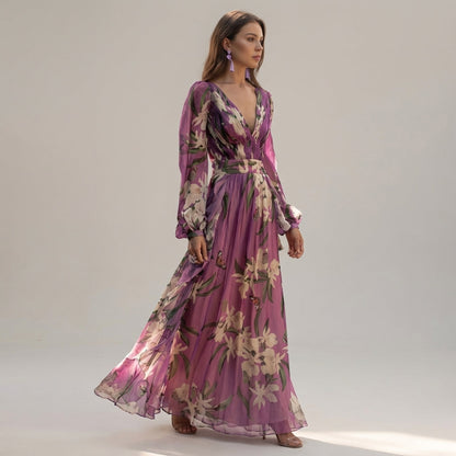 A woman stands wearing a floor-length lavender maxi dress made of sheer floral and butterfly print chiffon, featuring a deep V-neckline, pleated bodice, and long blouson sleeves.