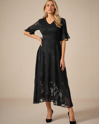 Black - A woman models a black formal midi dress featuring a V-neckline, flutter sleeves, and a textured floral jacquard overlay with a sheer hemline.