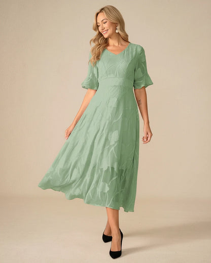 Green - A woman models a black formal midi dress featuring a V-neckline, flutter sleeves, and a textured floral jacquard overlay with a sheer hemline.