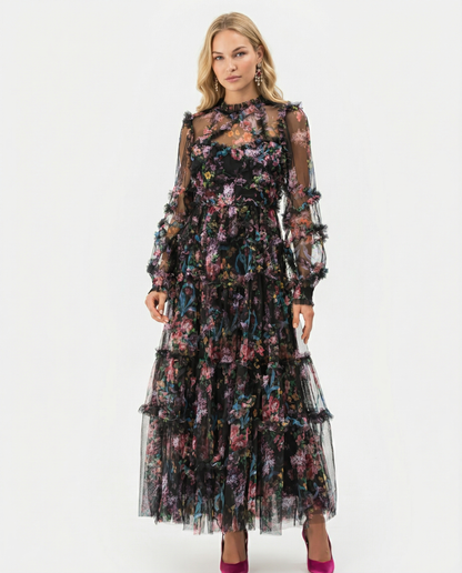 Black - A model wearing a high-neck, long-sleeved maxi dress in black tulle with a vibrant multi-coloured floral print, featuring tiered ruffles and sheer sleeves.