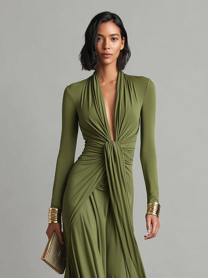 Green - A woman wears an olive green long-sleeve maxi dress crafted from fluid jersey, featuring a deep plunging V-neckline, a central ruched knot at the waist, and a cascading floor-length skirt.