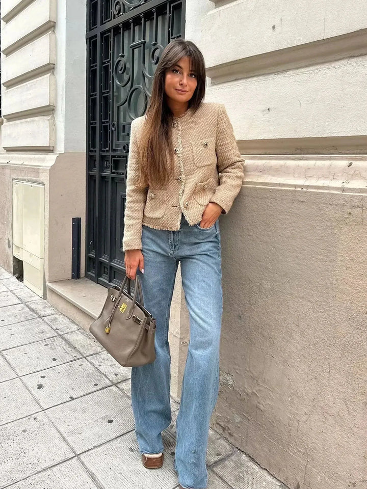 Beige - A woman wearing a beige textured boucle cropped jacket with gold buttons and fringed edges, styled over a light blue ruffled blouse and high-waisted jeans.
