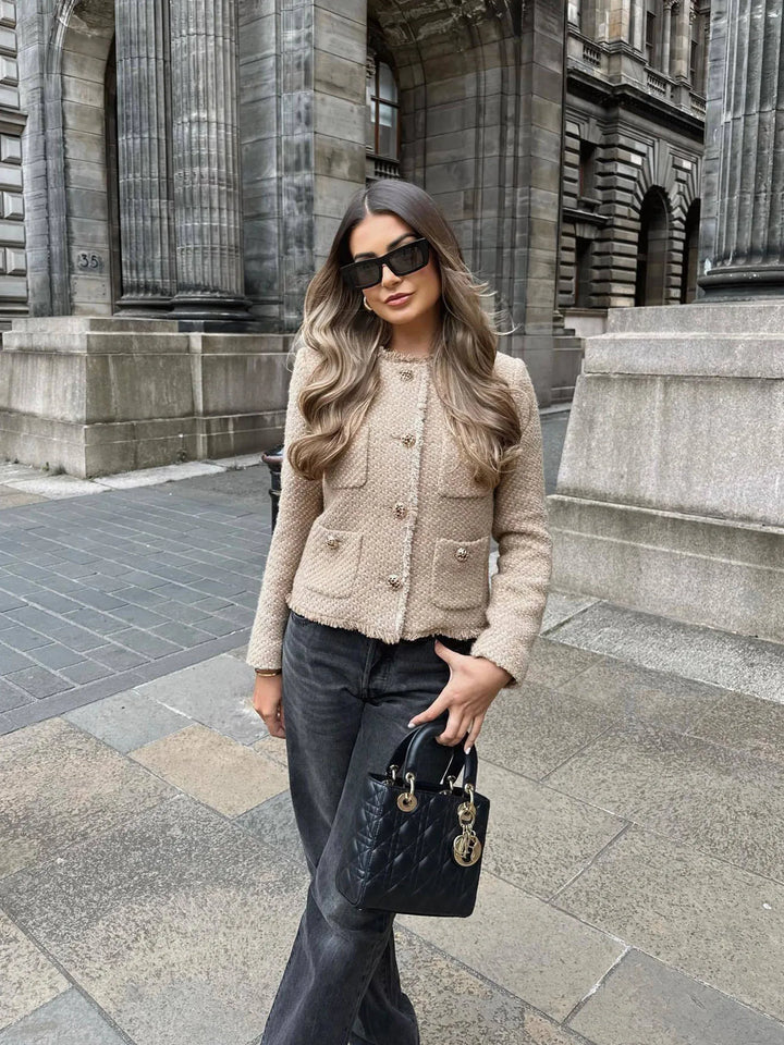 Beige - A woman wearing a beige textured boucle cropped jacket with gold buttons and fringed edges, styled over a light blue ruffled blouse and high-waisted jeans.