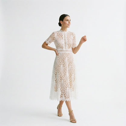 White - A woman wearing a black geometric lace midi dress with a high neck, short sleeves, and a defined waist, featuring a nude lining and scalloped hem.