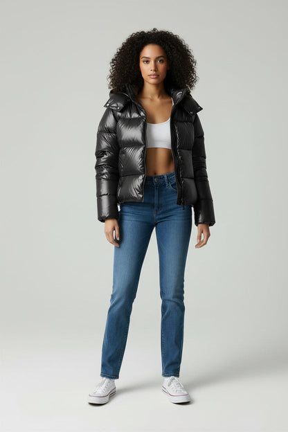 A woman wearing a glossy black hooded puffer jacket, a white crop top, blue high-waisted jeans, and white sneakers against a grey studio background.