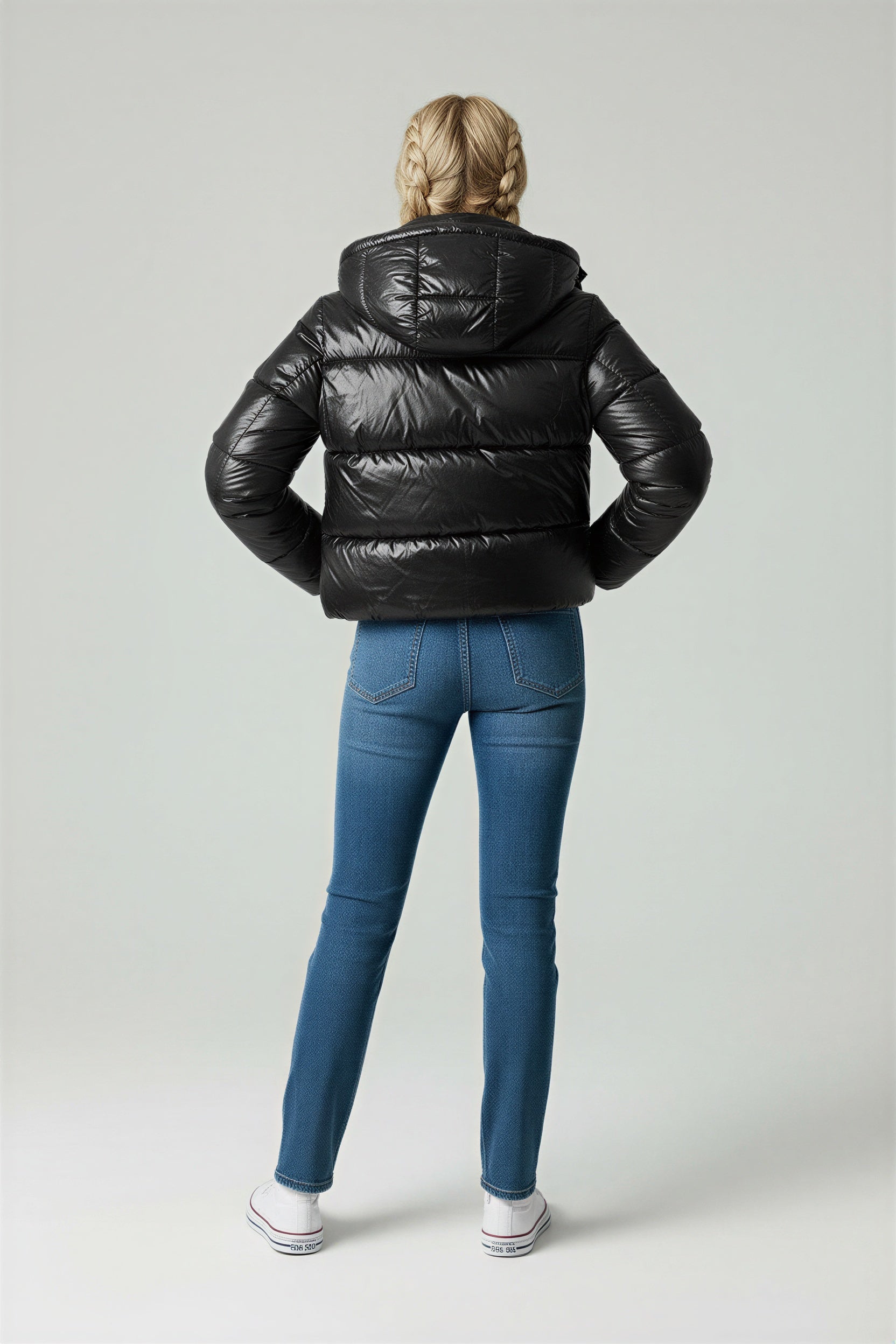 A woman wearing a glossy black hooded puffer jacket, a white crop top, blue high-waisted jeans, and white sneakers against a grey studio background.