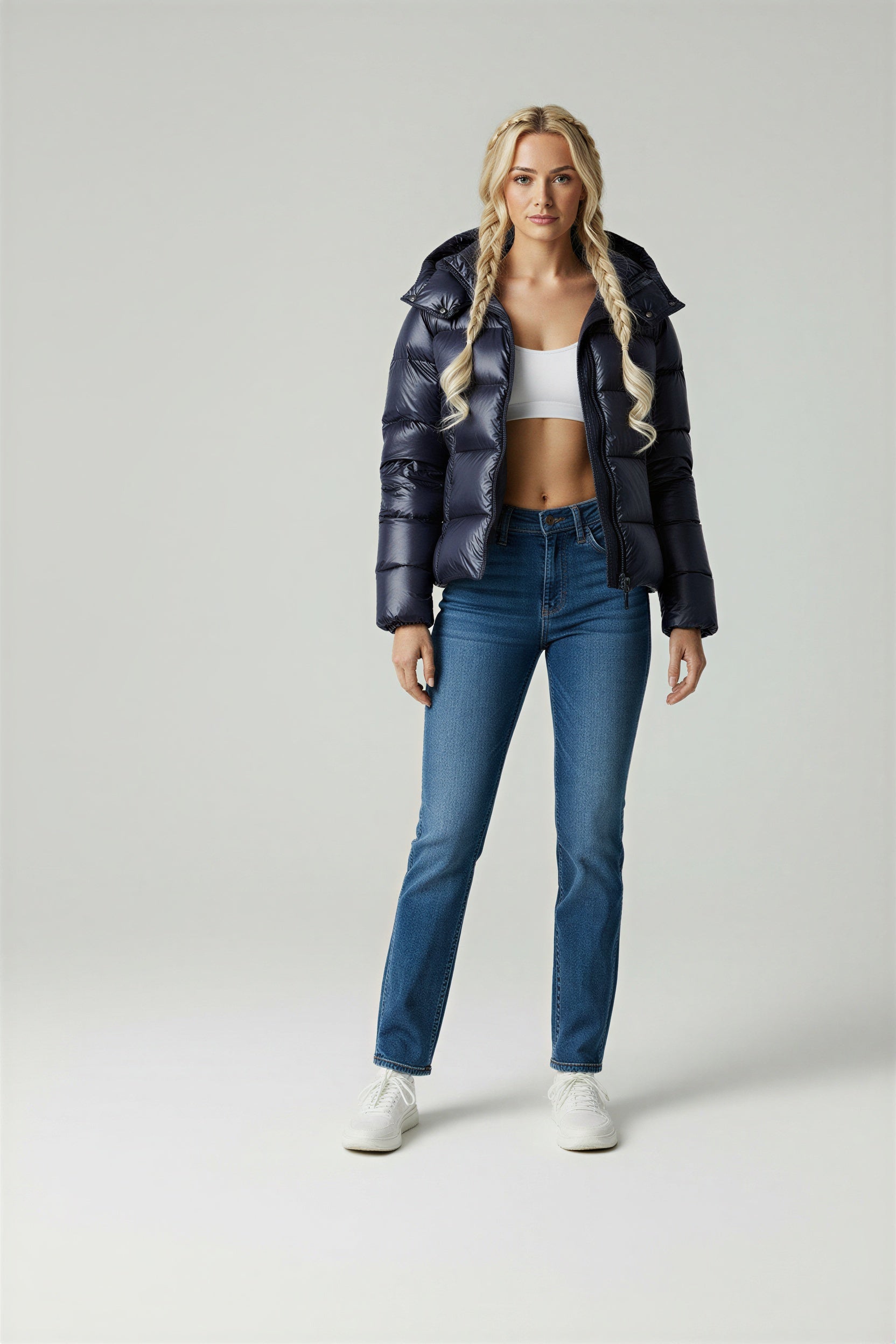 A woman wearing a glossy black hooded puffer jacket, a white crop top, blue high-waisted jeans, and white sneakers against a grey studio background.
