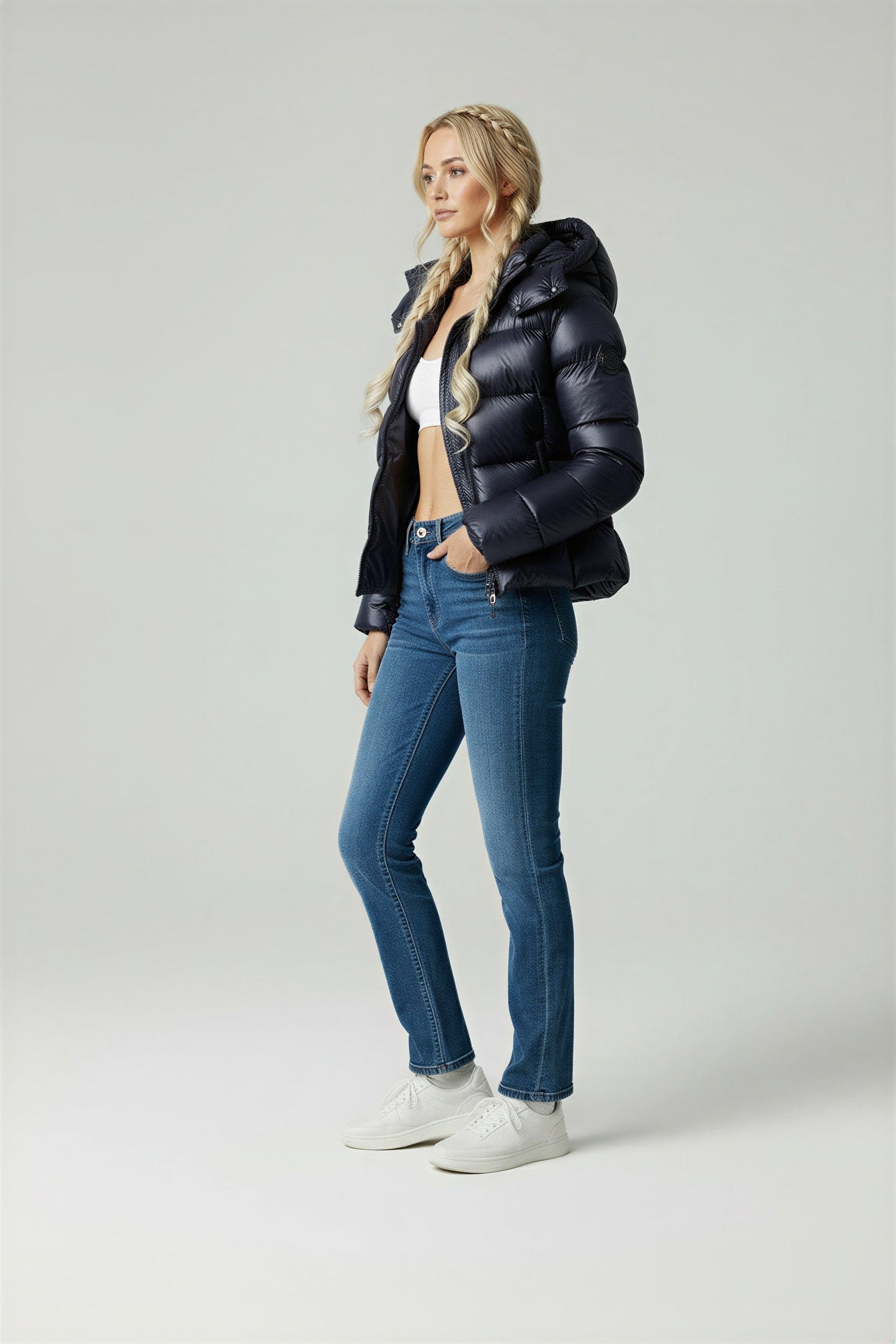 A woman wearing a glossy black hooded puffer jacket, a white crop top, blue high-waisted jeans, and white sneakers against a grey studio background.