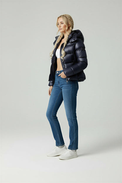 A woman wearing a glossy black hooded puffer jacket, a white crop top, blue high-waisted jeans, and white sneakers against a grey studio background.