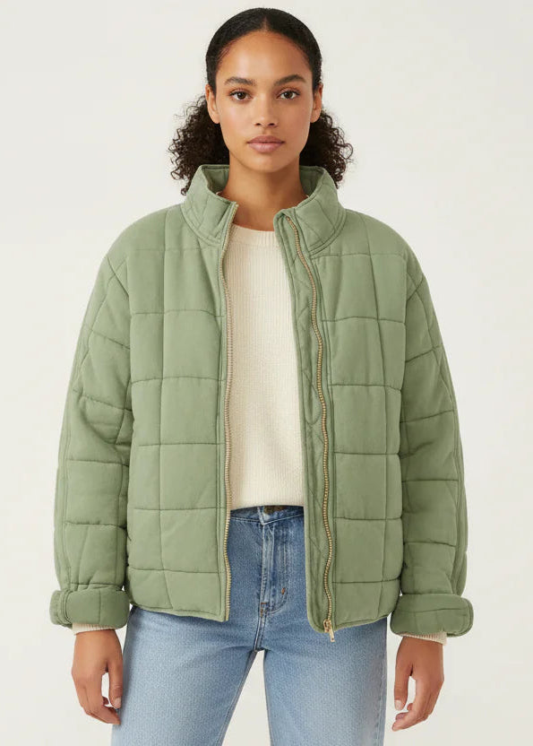 Green - A woman wearing a sage green grid-quilted jacket with a high funnel neck and gold-tone zip front, layered over a cream knit sweater and light-wash denim jeans.