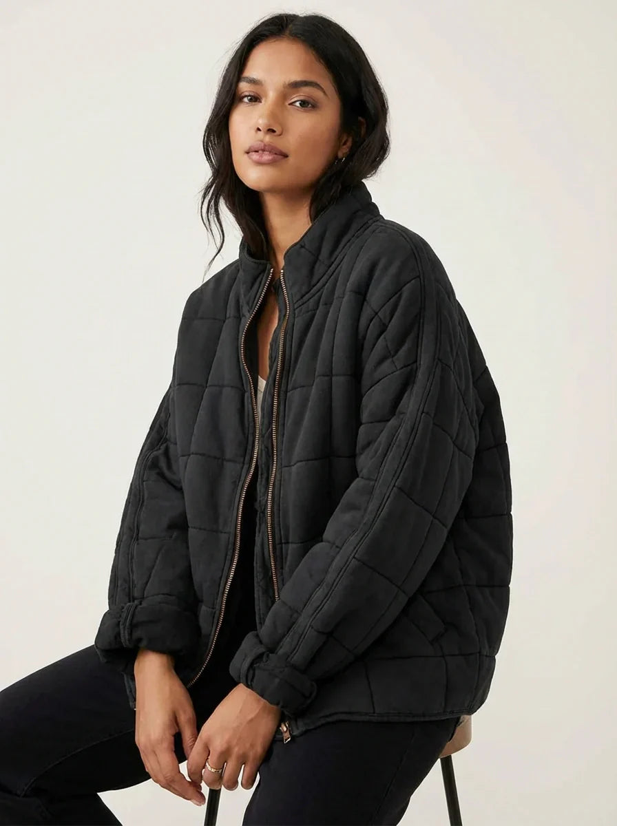 Black - A woman wearing a sage green grid-quilted jacket with a high funnel neck and gold-tone zip front, layered over a cream knit sweater and light-wash denim jeans.