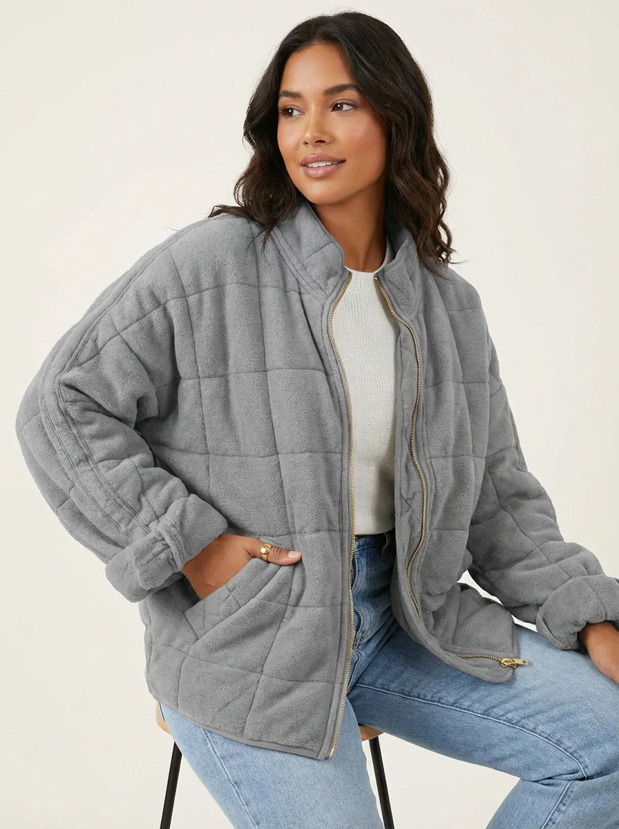Grey - A woman wearing a sage green grid-quilted jacket with a high funnel neck and gold-tone zip front, layered over a cream knit sweater and light-wash denim jeans.