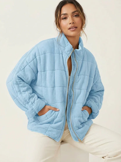 Light Blue - A woman wearing a sage green grid-quilted jacket with a high funnel neck and gold-tone zip front, layered over a cream knit sweater and light-wash denim jeans.