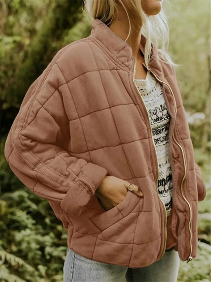 Brown - A woman wearing a sage green grid-quilted jacket with a high funnel neck and gold-tone zip front, layered over a cream knit sweater and light-wash denim jeans.