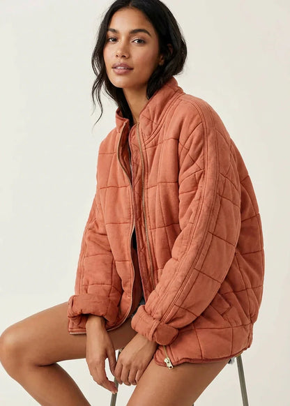 Orange - A woman wearing a sage green grid-quilted jacket with a high funnel neck and gold-tone zip front, layered over a cream knit sweater and light-wash denim jeans.