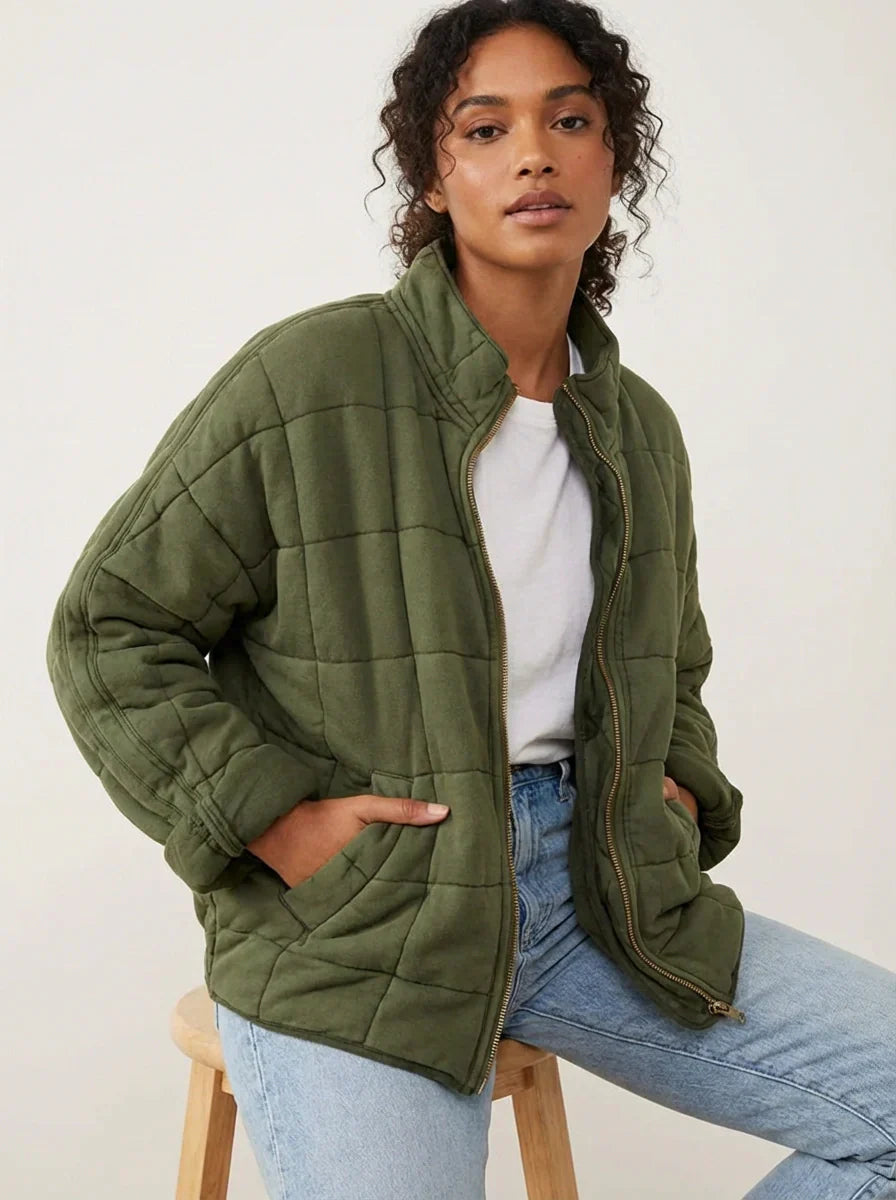 Olive Green - A woman wearing a sage green grid-quilted jacket with a high funnel neck and gold-tone zip front, layered over a cream knit sweater and light-wash denim jeans.