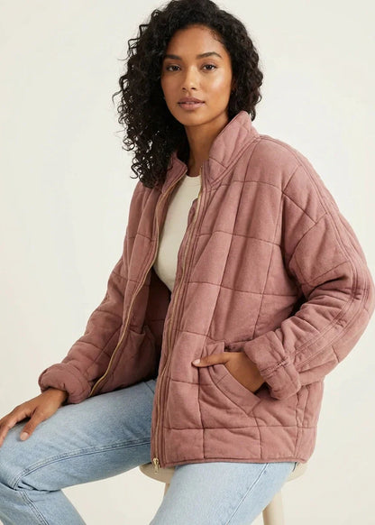 Olive Green - A woman wearing a sage green grid-quilted jacket with a high funnel neck and gold-tone zip front, layered over a cream knit sweater and light-wash denim jeans.