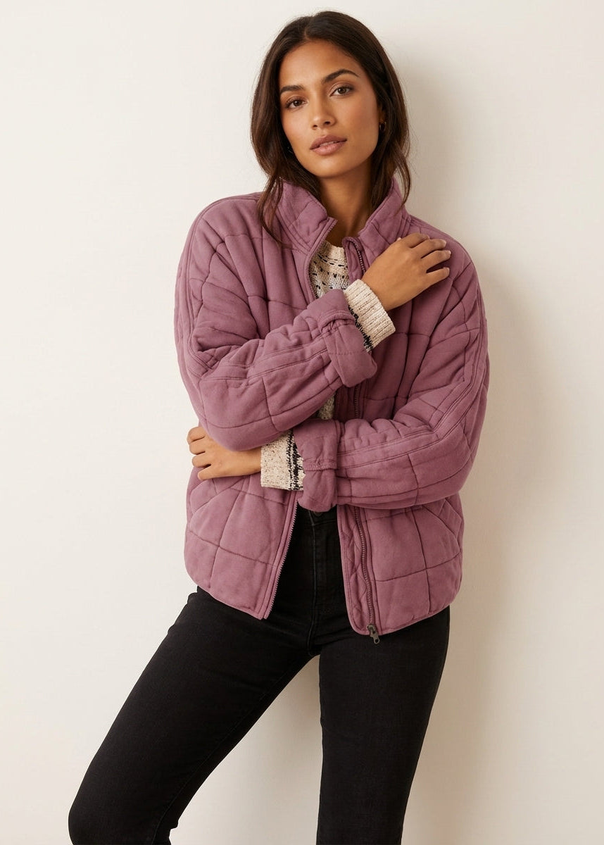 Pink - A woman wearing a sage green grid-quilted jacket with a high funnel neck and gold-tone zip front, layered over a cream knit sweater and light-wash denim jeans.