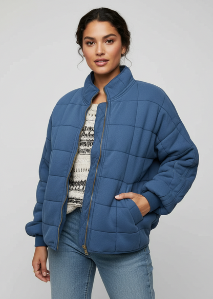 Blue - A woman wearing a sage green grid-quilted jacket with a high funnel neck and gold-tone zip front, layered over a cream knit sweater and light-wash denim jeans.