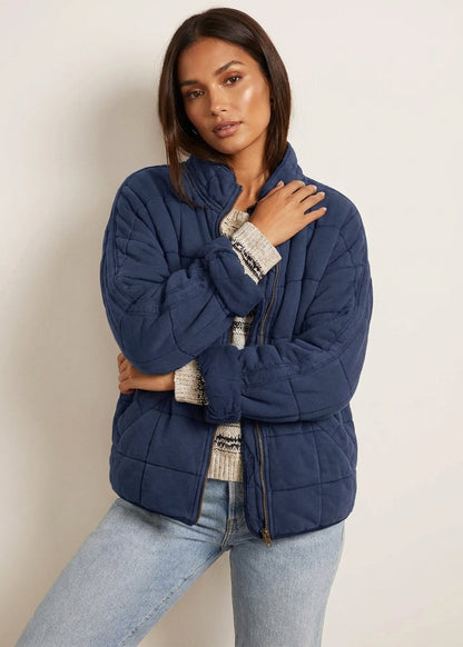Navy Blue - A woman wearing a sage green grid-quilted jacket with a high funnel neck and gold-tone zip front, layered over a cream knit sweater and light-wash denim jeans.