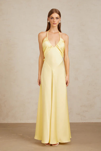 A woman wearing a pale yellow, formal halter neck satin maxi dress with a ruched bust and flowing silhouette against a neutral background.