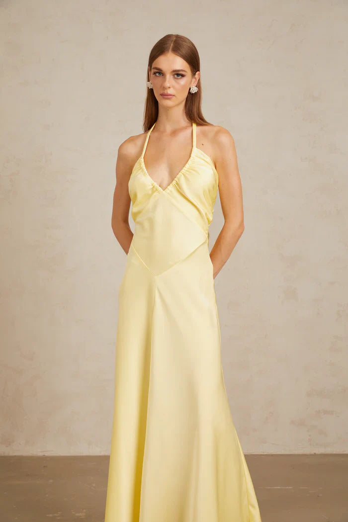 A woman wearing a pale yellow, formal halter neck satin maxi dress with a ruched bust and flowing silhouette against a neutral background.