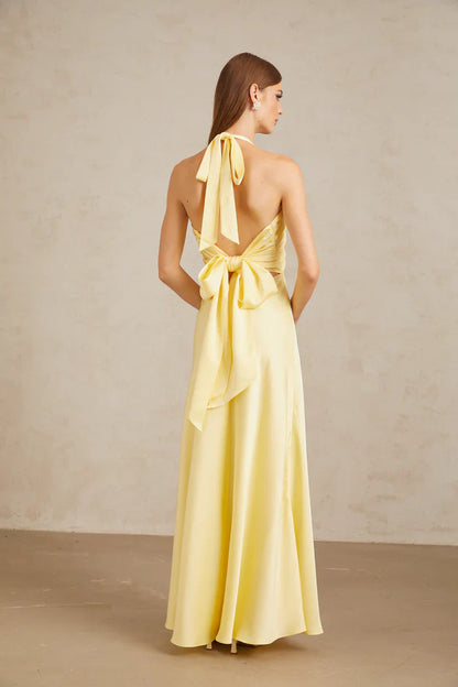 A woman wearing a pale yellow, formal halter neck satin maxi dress with a ruched bust and flowing silhouette against a neutral background.