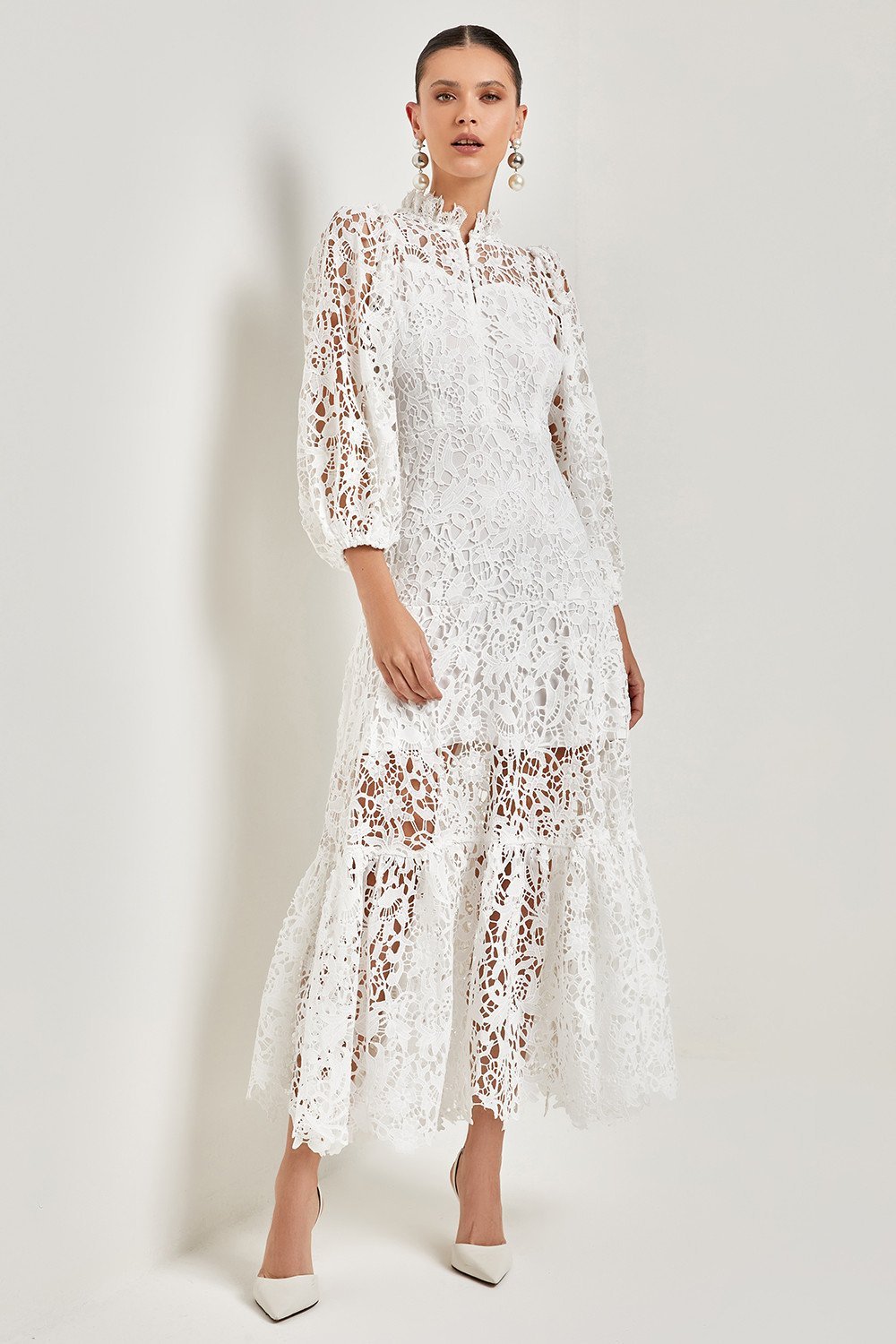White - A woman models a vibrant green, long-sleeve maxi dress featuring an intricate floral lace overlay, a high neckline, and a tiered skirt, holding a matching green clutch in a greenhouse setting.