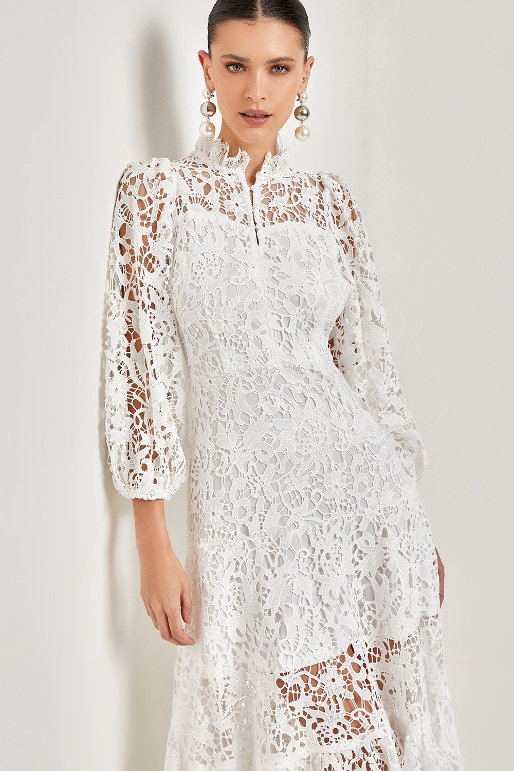 White - A woman models a vibrant green, long-sleeve maxi dress featuring an intricate floral lace overlay, a high neckline, and a tiered skirt, holding a matching green clutch in a greenhouse setting.