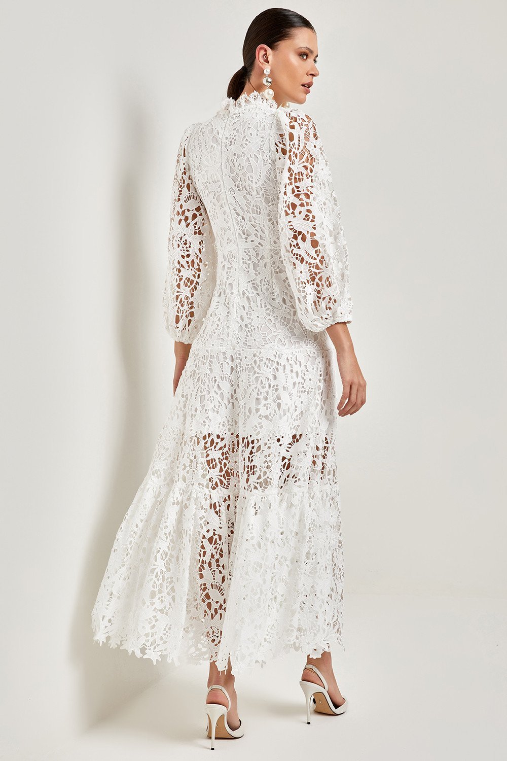 White - A woman models a vibrant green, long-sleeve maxi dress featuring an intricate floral lace overlay, a high neckline, and a tiered skirt, holding a matching green clutch in a greenhouse setting.