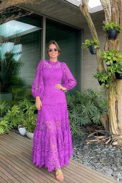 Purple - A woman models a vibrant green, long-sleeve maxi dress featuring an intricate floral lace overlay, a high neckline, and a tiered skirt, holding a matching green clutch in a greenhouse setting.