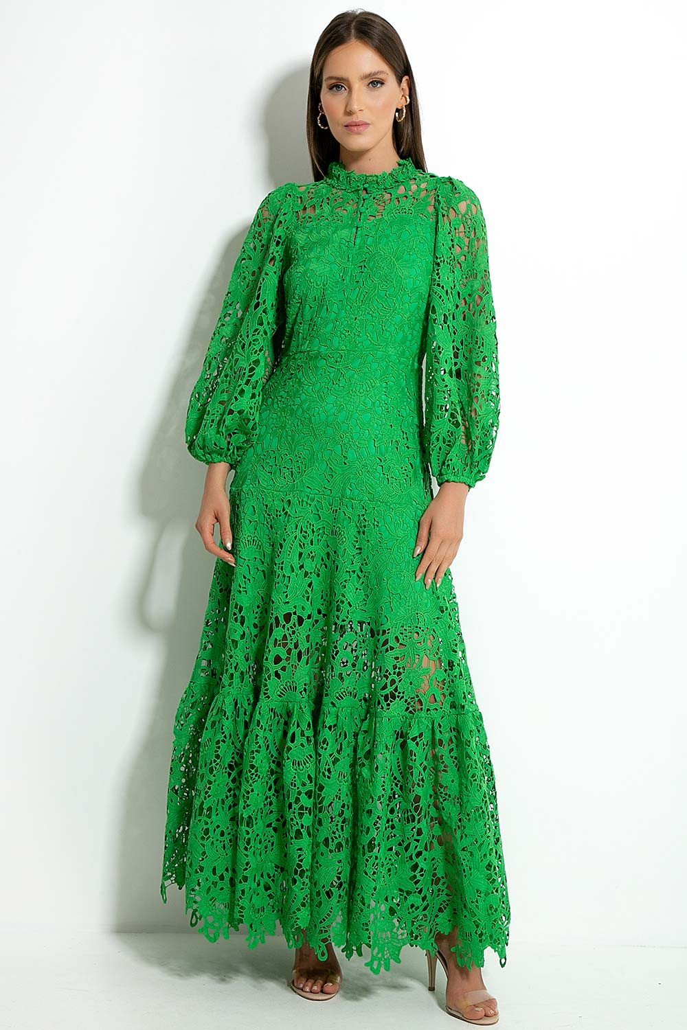Green - A woman models a vibrant green, long-sleeve maxi dress featuring an intricate floral lace overlay, a high neckline, and a tiered skirt, holding a matching green clutch in a greenhouse setting.